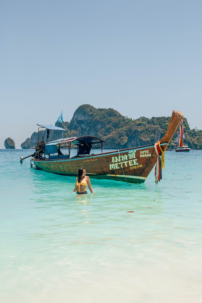A woman wades into crystal clear water near a traditional longtail boat in Thailand.