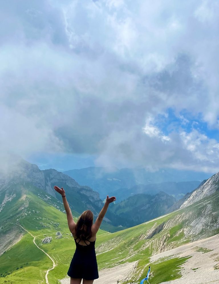 a girl stands with her back facing the camera looking out on mountains with her arms raised above her head