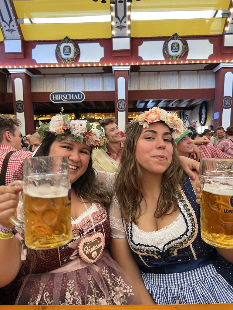 two girls wearing drindls hold beers and smile to the camera while sitting at a table at Oktoberfest