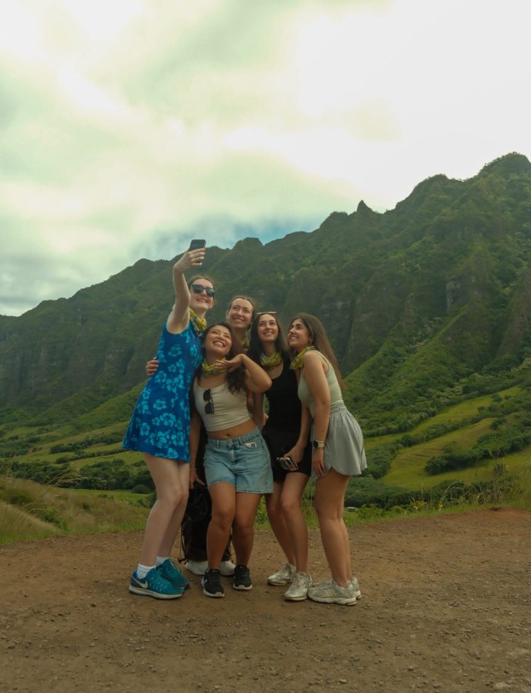 A group of girls poses for a selfie in front of the rolling green hills of Kualoa ranch on Oahu