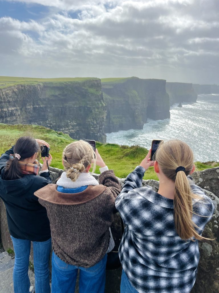 Three girls face away from the camera taking pictures of the Cliffs of Moher from the top walkways of the cliffs