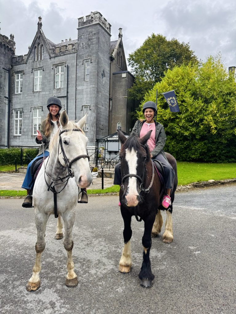 two girls atop horses pause for a picture in front of a stone castle in Ireland