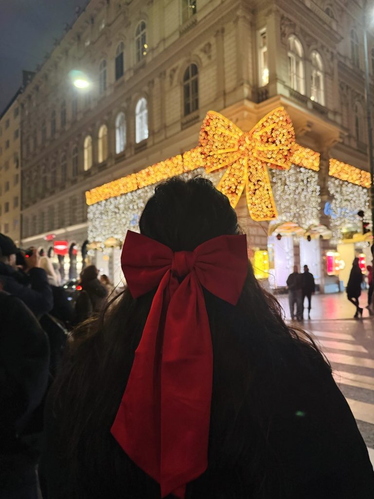 a girl faces away from the camera wearing a big red bow in her hair, looking at a light up bow Christmas decoration in Vienna