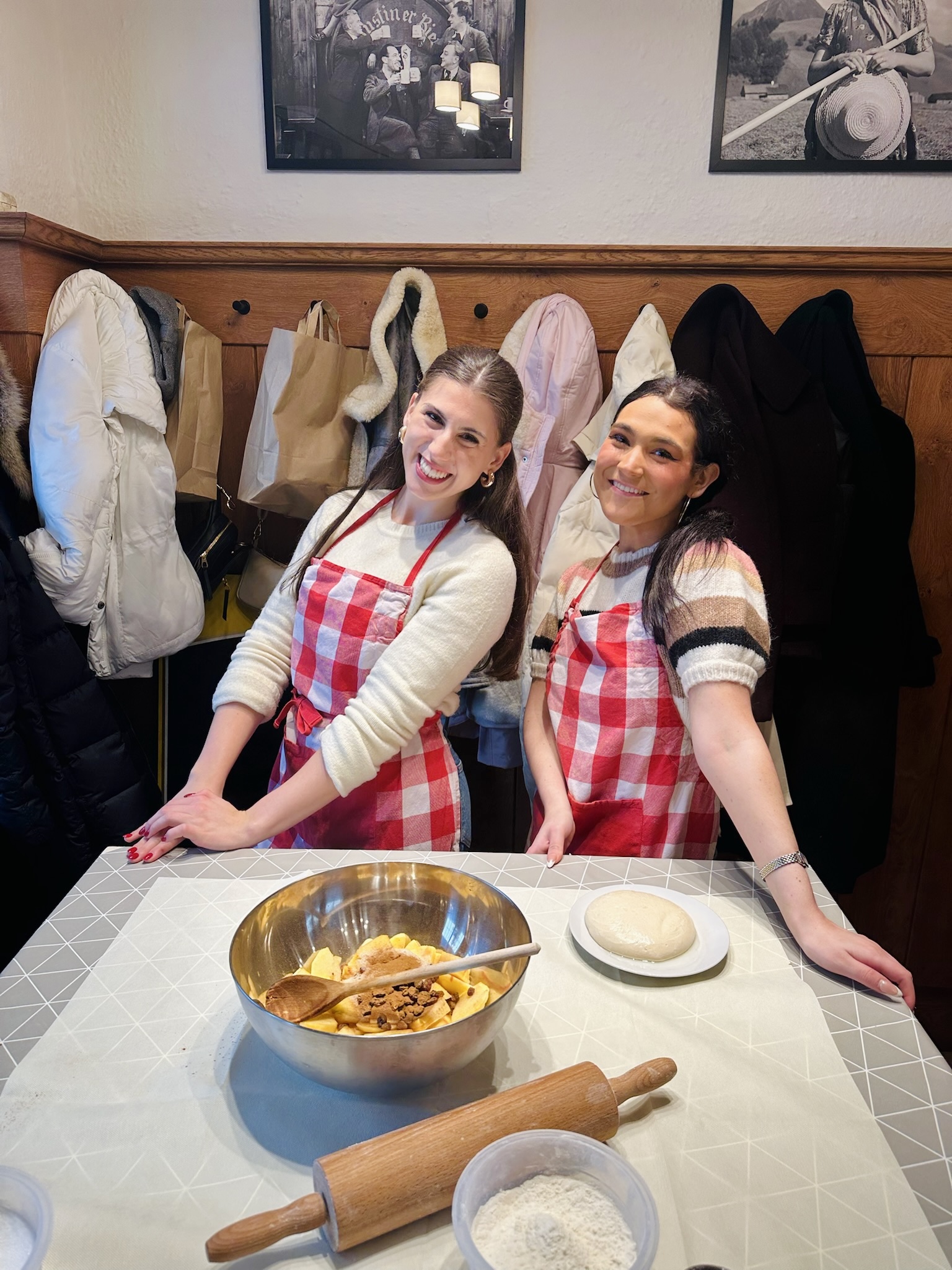 two girls dressed in white sweaters and red plaid aprons pose for a picture behind a table with apples and a rolling pin for a cooking class