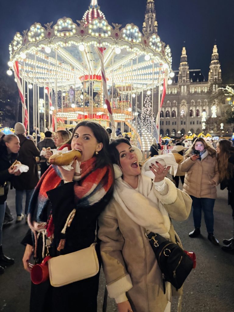 two girls pretend to eat bratwursts at a Christmas market in Vienna lit with lights