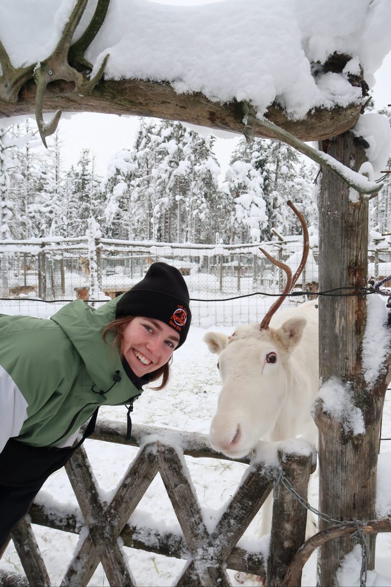 girl bundled up in winter gear poses smiling next to a white reindeer in a wooden corral