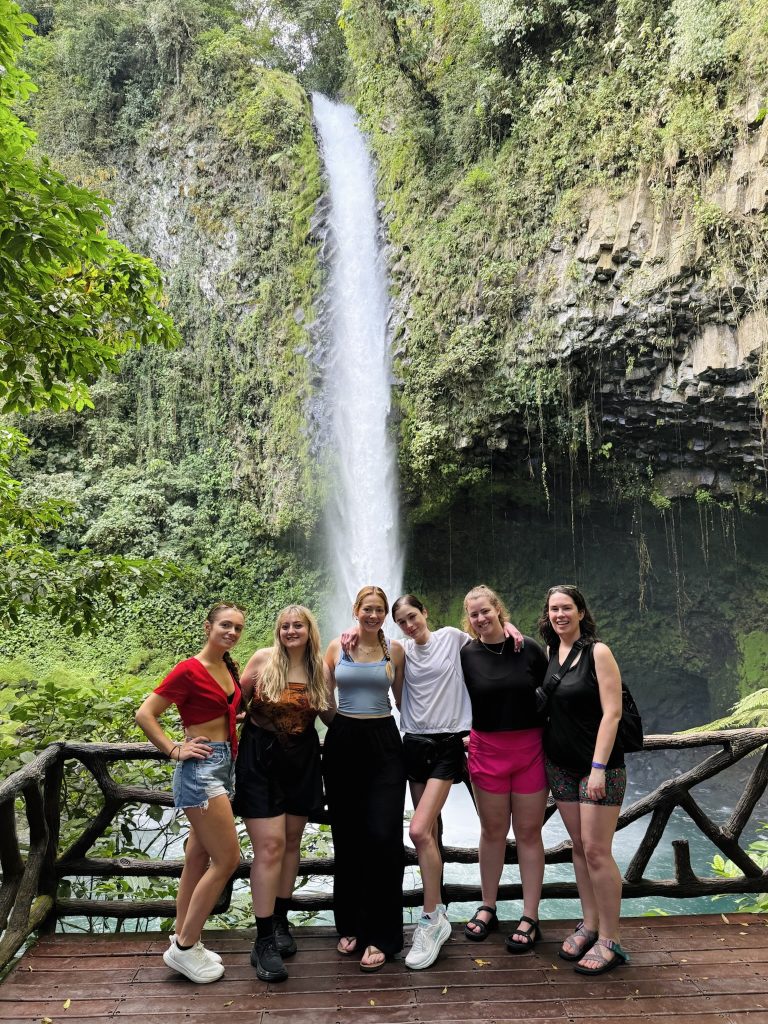 six women pose smiling on a wooden platform in front of La Fortuna waterfall in the Costa Rica rainforest